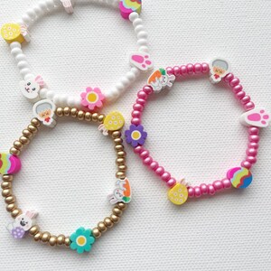 May include: Three beaded bracelets with Easter-themed charms. The bracelets are white, pink, and gold. The charms include bunnies, flowers, eggs, and footprints.