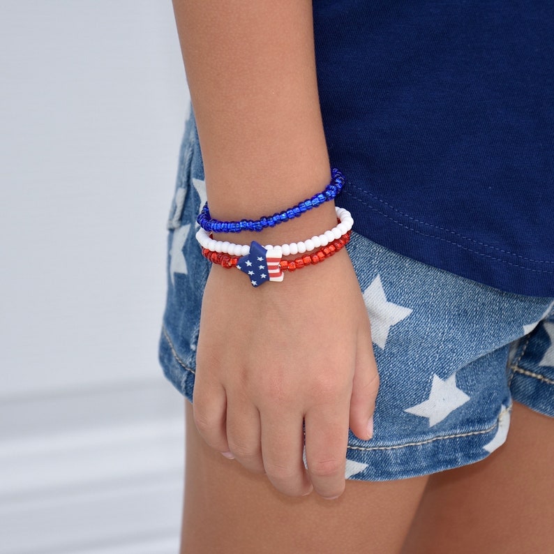 May include: A child's wrist wearing three beaded bracelets. The bracelets are red, white, and blue, and feature a small American flag charm.
