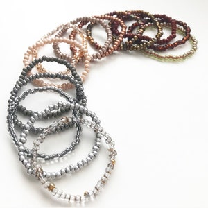 May include: A set of 14 beaded bracelets in various shades of brown, gray, and silver. The bracelets are arranged in a semi-circle, with the darkest shades at the top and the lightest shades at the bottom.