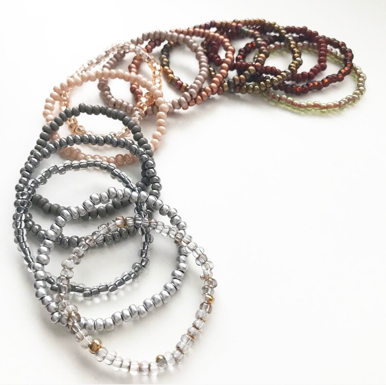 May include: A set of 14 beaded bracelets in various shades of brown, gray, and silver. The bracelets are arranged in a semi-circle, with the darkest shades at the top and the lightest shades at the bottom.