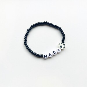 K&ouml;nnte beinhalten: Schwarzes Perlenarmband mit wei&szlig;en Buchstabenperlen, die "MACY" buchstabieren, und einem wei&szlig;en Pfotenabdruck-Anh&auml;nger.