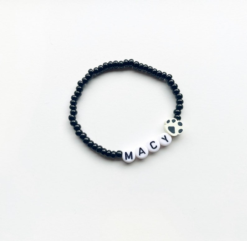 K&ouml;nnte beinhalten: Schwarzes Perlenarmband mit wei&szlig;en Buchstabenperlen, die "MACY" buchstabieren, und einem wei&szlig;en Pfotenabdruck-Anh&auml;nger.