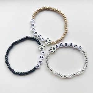 K&ouml;nnte beinhalten: Drei Perlenarmb&auml;nder mit Pfotenanh&auml;ngern. Die Armb&auml;nder sind schwarz, gold und klar. Die Armb&auml;nder buchstabieren die Namen "Macy", "Cooper" und "Speedy".