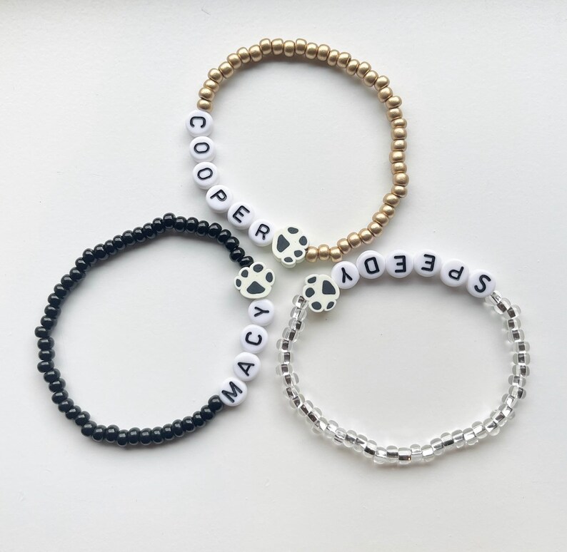 K&ouml;nnte beinhalten: Drei Perlenarmb&auml;nder mit Pfotenanh&auml;ngern. Die Armb&auml;nder sind schwarz, gold und klar. Die Armb&auml;nder buchstabieren die Namen "Macy", "Cooper" und "Speedy".