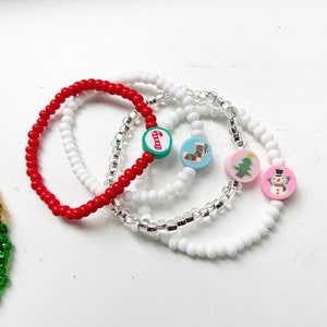 Puede incluir: Cuatro pulseras con tem&aacute;tica navide&ntilde;a con diferentes dijes. Una pulsera es roja, una es blanca, una es transparente con cuentas plateadas y una es blanca con cuentas plateadas. Los dijes incluyen una ca&ntilde;a de az&uacute;car, una media, un &aacute;rbol de Navidad y un mu&ntilde;eco de nieve.