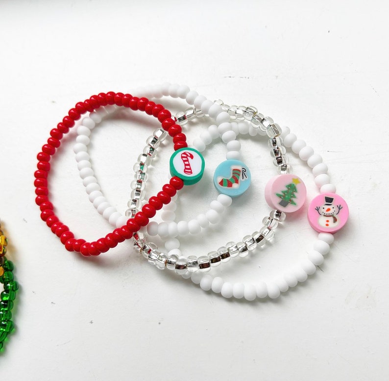 Puede incluir: Cuatro pulseras con tem&aacute;tica navide&ntilde;a con diferentes dijes. Una pulsera es roja, una es blanca, una es transparente con cuentas plateadas y una es blanca con cuentas plateadas. Los dijes incluyen una ca&ntilde;a de az&uacute;car, una media, un &aacute;rbol de Navidad y un mu&ntilde;eco de nieve.