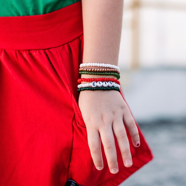 Puede incluir: Primer plano de la mu&ntilde;eca de una persona que lleva varias pulseras de cuentas de colores, incluida una pulsera de cuentas blancas con el nombre "ELISE" escrito en negro.