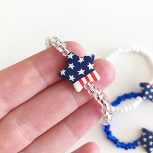 May include: A beaded bracelet with a star-shaped charm featuring the American flag. The bracelet is made with clear beads and the star charm is red, white, and blue.