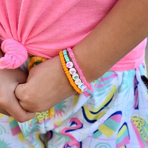 May include: A child's wrist with four bracelets. The top bracelet is pink and clear. The next bracelet is light blue and clear. The third bracelet is orange and has the name "HARPER" spelled out in colorful beads. The bottom bracelet is pink and clear.