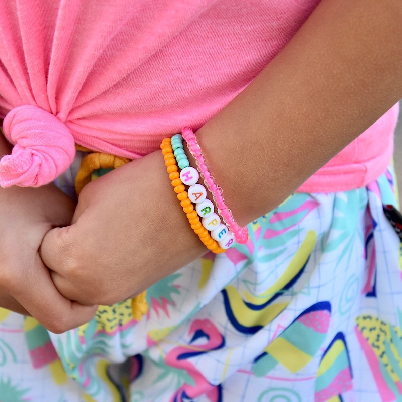 May include: A child's wrist with four bracelets. The top bracelet is pink and clear. The next bracelet is light blue and clear. The third bracelet is orange and has the name "HARPER" spelled out in colorful beads. The bottom bracelet is pink and clear.