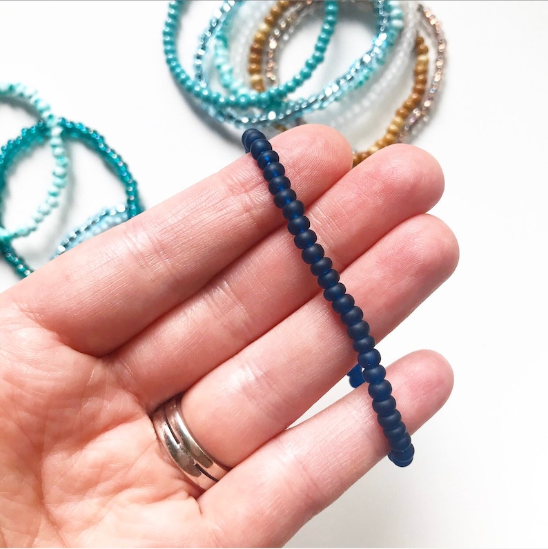 May include: A dark blue beaded bracelet made of small, round beads. The bracelet is shown on a hand, with the beads forming a simple, elegant design.