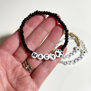 K&ouml;nnte beinhalten: Schwarzes Perlenarmband mit wei&szlig;en Buchstabenperlen, die "MACY" buchstabieren, und einem Pfotenanh&auml;nger.
