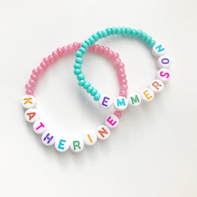 May include: Two beaded bracelets with the names "Katherine" and "Emmerson" spelled out in colorful letters on white beads. The bracelets are pink and turquoise.