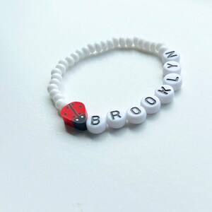 K&ouml;nnte beinhalten: Wei&szlig;es Perlenarmband mit dem Namen "BROOKLYN" in schwarzen Buchstaben. Ein roter und schwarzer Marienk&auml;fer-Anh&auml;nger ist am Armband befestigt.
