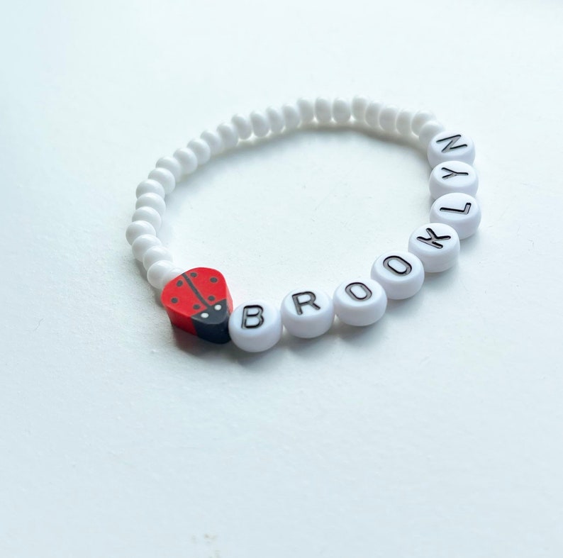 K&ouml;nnte beinhalten: Wei&szlig;es Perlenarmband mit dem Namen "BROOKLYN" in schwarzen Buchstaben. Ein roter und schwarzer Marienk&auml;fer-Anh&auml;nger ist am Armband befestigt.