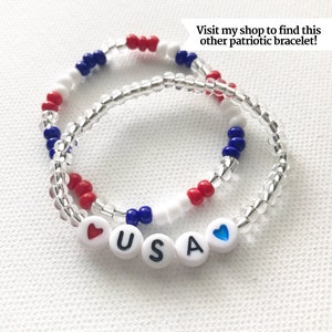 May include: A red, white, and blue beaded bracelet with the word "USA" spelled out in white beads with a red heart bead on one end and a blue heart bead on the other end.