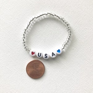 May include: A white beaded bracelet with the letters "USA" and red and blue hearts. The bracelet is next to a US penny.