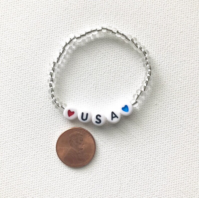 May include: A white beaded bracelet with the letters "USA" and red and blue hearts. The bracelet is next to a US penny.