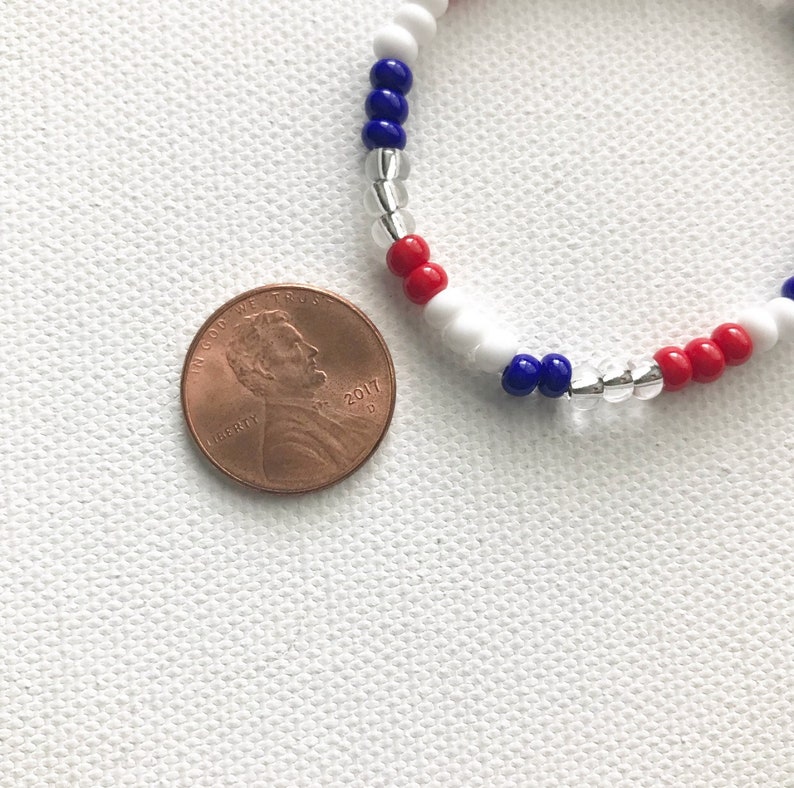 May include: A red, white, and blue beaded bracelet with silver beads. The bracelet is next to a US penny.