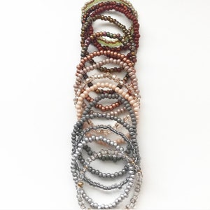 May include: A set of 12 beaded bracelets in various shades of brown, beige, and gray. The bracelets are stacked on top of each other, creating a gradient effect.