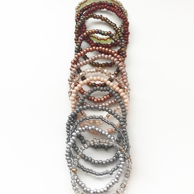May include: A set of 12 beaded bracelets in various shades of brown, beige, and gray. The bracelets are stacked on top of each other, creating a gradient effect.