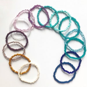 May include: A set of 11 colorful beaded bracelets in various shades of purple, blue, and brown. The bracelets are made of small, round beads and are arranged in a circle.