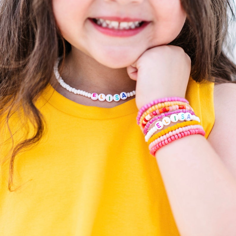 May include: A young girl wearing a yellow shirt with a white beaded necklace that spells "ELISA". She is also wearing a stack of colorful beaded bracelets that spell "ELISA" on one of the bracelets.