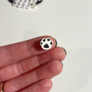 K&ouml;nnte beinhalten: Ein kleiner wei&szlig;er Keramik-Pfotenabdruck-Anh&auml;nger mit schwarzen Details. Der Anh&auml;nger wird in einer Hand gehalten.