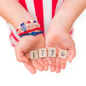 May include: A child's hands holding up wooden blocks with the numbers 1776 written on them. The child is wearing red, white, and blue bracelets.