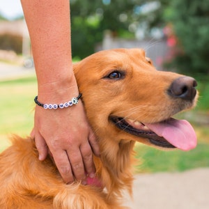 K&ouml;nnte beinhalten: Eine Person tr&auml;gt ein schwarzes Armband mit wei&szlig;en Perlen, die "DOG MOM" buchstabieren, und einem Pfotenanh&auml;nger und streichelt einen Golden Retriever.