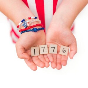 May include: A child's hands holding up four wooden blocks with the numbers 1776 written on them. The child is wearing red, white, and blue bracelets.