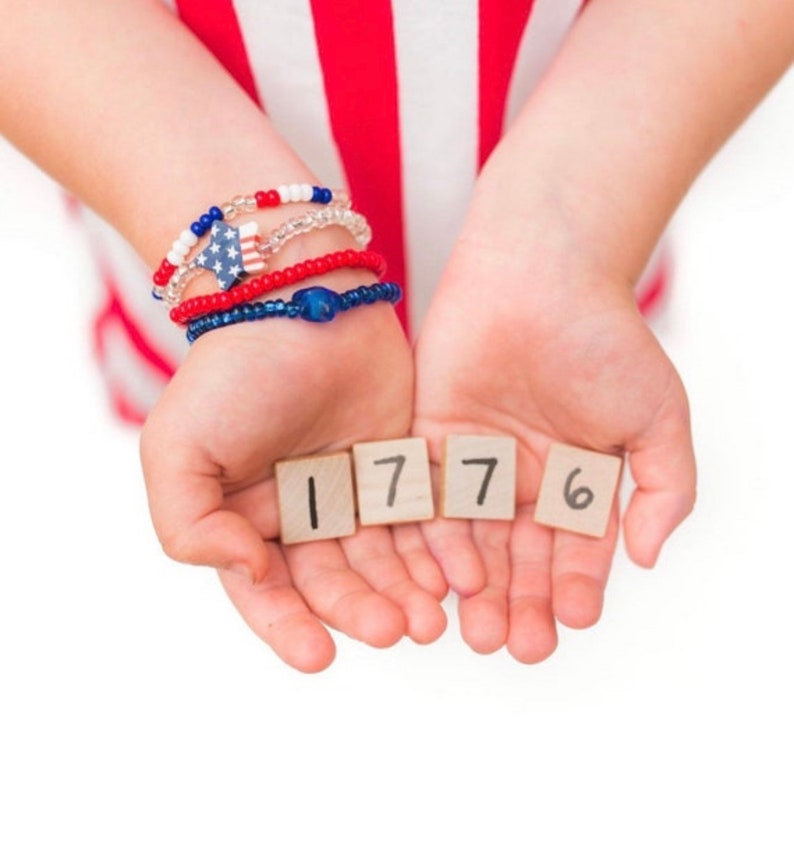 May include: A child's hands holding up four wooden blocks with the numbers 1776 written on them. The child is wearing red, white, and blue bracelets.