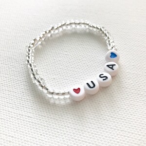 May include: A white beaded bracelet with the letters "USA" and two hearts, one red and one blue.