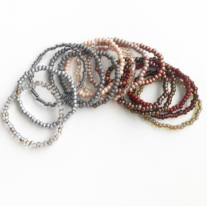 May include: A set of 12 beaded bracelets in various shades of gray, brown, and beige. The bracelets are stacked on top of each other, creating a gradient effect.