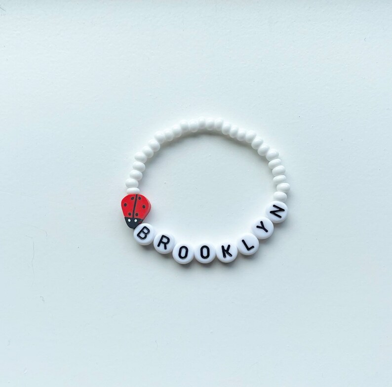 K&ouml;nnte beinhalten: Wei&szlig;es Perlenarmband mit dem Namen "BROOKLYN" in schwarzen Buchstaben. Ein roter Marienk&auml;fer-Anh&auml;nger ist am Armband befestigt.