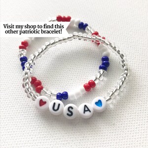 May include: Two beaded bracelets with red, white, and blue beads. The bracelets are layered and spell out "USA" with a red heart on one side and a blue heart on the other.