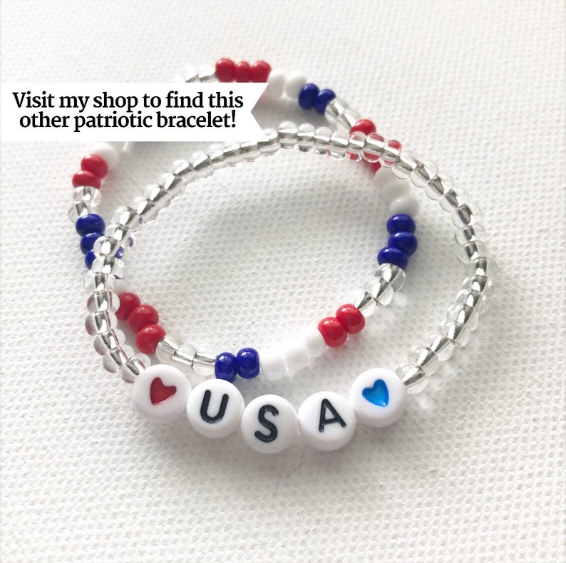 May include: Two beaded bracelets with red, white, and blue beads. The bracelets are layered and spell out "USA" with a red heart on one side and a blue heart on the other.