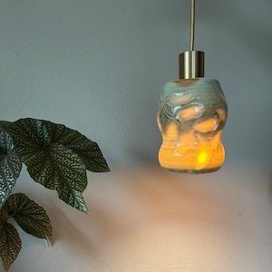 May include: A ceramic pendant light fitting with a light green glaze and a gold metal hanging cord. The light is on and casting a warm glow.