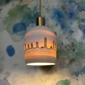 May include: A white ceramic pendant light with a gold metal top and a green glaze dripping down the sides. The light is hanging from a ceiling.