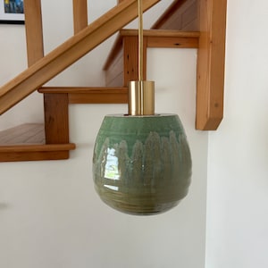 Puede incluir: Una lámpara colgante de cerámica verde con una fijación de metal dorado. La luz cuelga de un cable y está delante de una escalera de madera.
