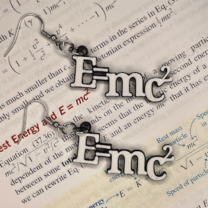 May include: A pair of silver earrings with a white enamel charm that says "E=mc²". The earrings are hanging from a book with the equation "E=mc²" printed on it.