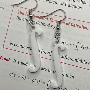 May include: A pair of silver earrings with white, 3D printed charms shaped like the integral symbol. The charms are attached to the earrings with silver hooks. The earrings are displayed on a page of a math textbook that includes the text "The Fundamental Theorem of Calculus".