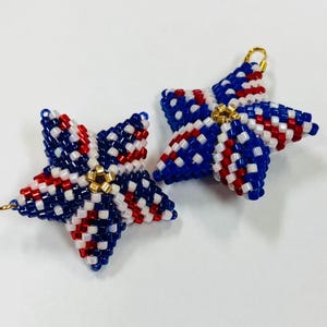 May include: Two beaded star ornaments in red, white, and blue. The stars have a woven pattern and a gold-colored loop at the top for hanging. The ornaments are likely handmade and suitable for patriotic or holiday decor.