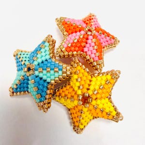 May include: Four beaded star ornaments in shades of blue, pink, orange, and yellow. Each star is outlined with gold-colored beads. The stars are arranged on a white surface.
