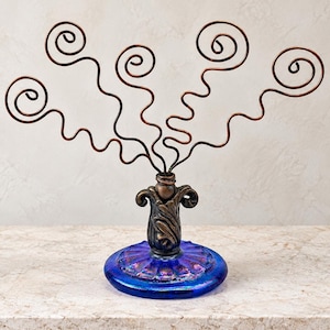 May include: A decorative display stand featuring four copper-colored wire stems with spiral tops. The stems emerge from a dark, ornate metal base with leaf-like detailing, which is mounted onto a circular, iridescent blue glass pedestal.