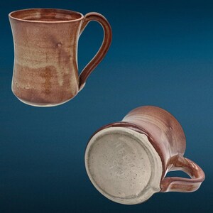 May include: Two ceramic mugs with a glossy, mottled brown and tan glaze. One mug is shown upright with a curved handle, while the other is positioned on its side to reveal the light-colored, speckled, unglazed circular base.