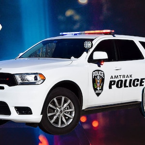 May include: A white Dodge Durango SUV with black accents and a blue and red light bar on the roof. The vehicle is marked "AMTRAK POLICE" with a gold and black badge on the side. The badge features an eagle and the words "AMTRAK POLICE".