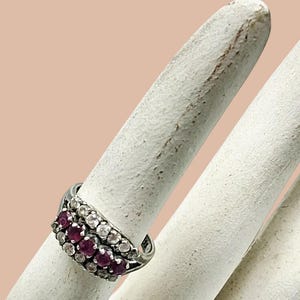May include: A silver-toned ring featuring three horizontal rows of small, round-cut stones. The center row consists of deep red stones, while the top and bottom rows are composed of clear, colorless stones set in a metallic band design.