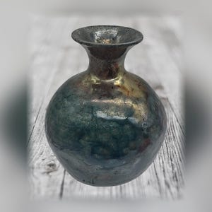 May include: A small, round ceramic vase with a narrow neck and a flared rim. The surface features a mottled, iridescent glaze with blended tones of deep blue, dark green, and metallic bronze, creating a textured, reflective appearance.