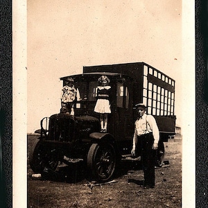 May include: Vintage black and white photograph of a truck with three children standing on it. A man stands beside the truck. The truck has large wheels and a boxy back with windows.